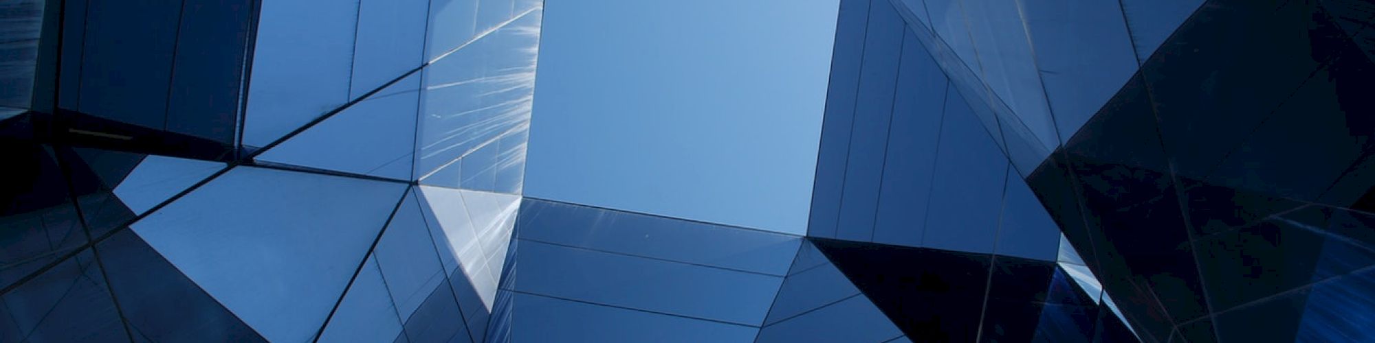 Looking up at a geometric structure made of reflective glass panels, creating a symmetrical and abstract view with a clear blue sky above.