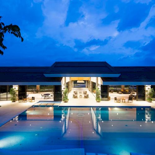 A luxurious, illuminated resort with a large, inviting swimming pool under a dusky blue sky.