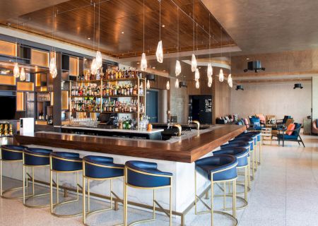 A modern bar with a wooden counter, blue cushioned stools, hanging lights, and shelves stocked with bottles, located in a stylish interior.