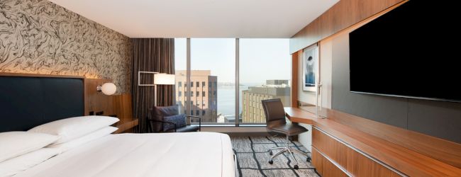 A modern hotel room with a large bed, wall-mounted TV, desk, chair, and a window showing a cityscape view.