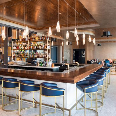 The image shows a modern bar with blue stools, a central counter, hanging lights, and a stocked bar area in an upscale setting.