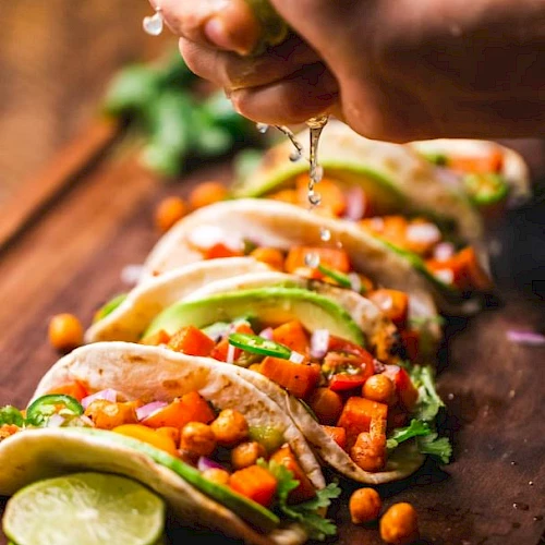 A hand squeezes lime over a row of tacos filled with vegetables and garnished with lime slices and greens on a wooden board.