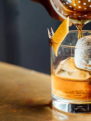 A drink is being poured into a glass with ice and a citrus twist garnish on a bar counter.