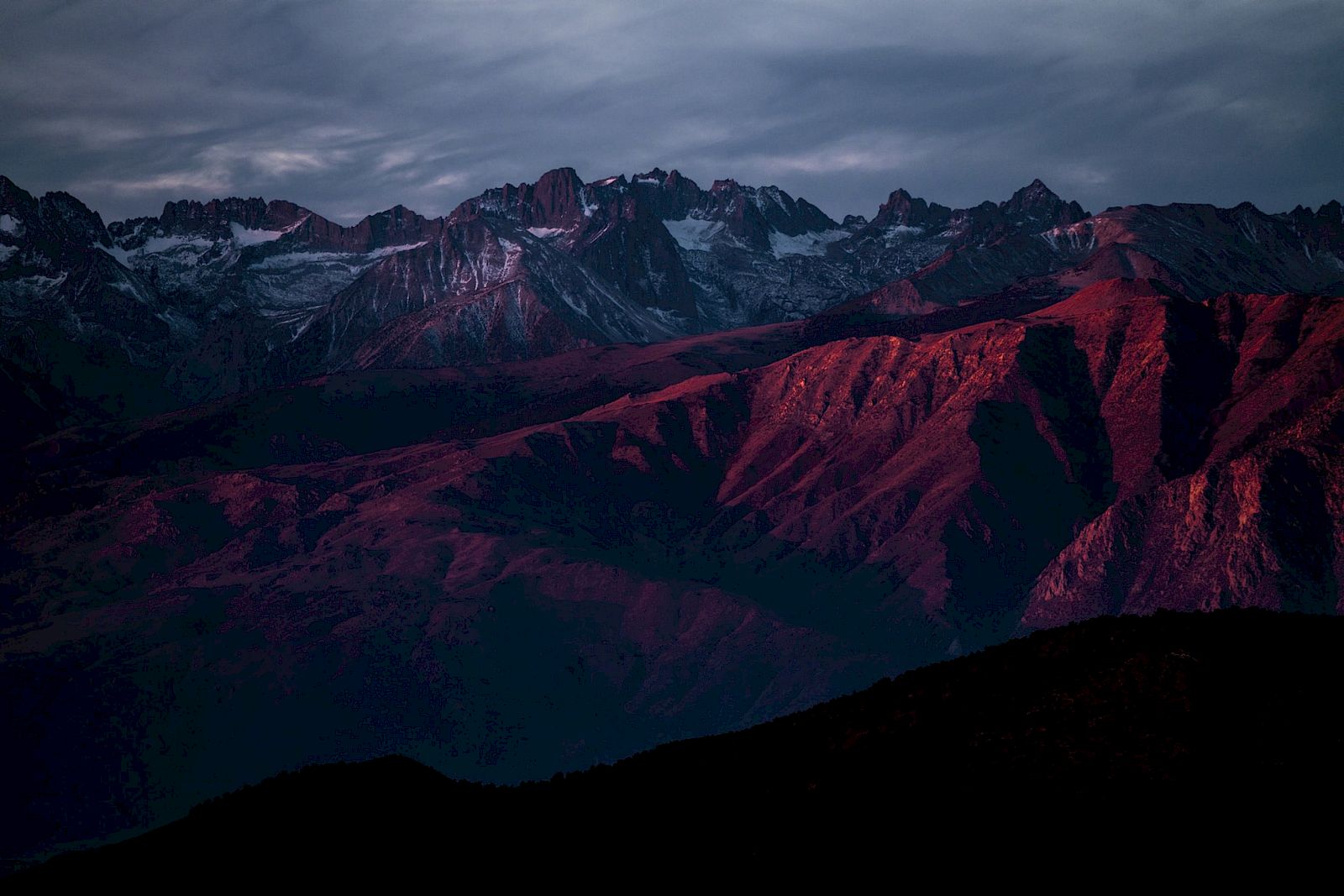 A mountain range at sunset, with peaks illuminated in red hues, under a moody, cloud-filled sky.