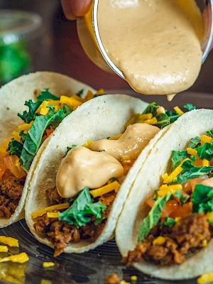 Three tacos filled with meat, cheese, and greens on a plate, with sauce being poured over them.