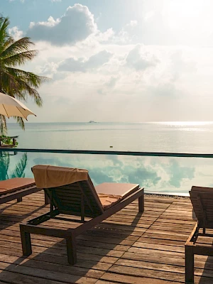 A serene poolside scene with lounge chairs, umbrellas, and palm trees overlooking a calm ocean under a partly cloudy sky.