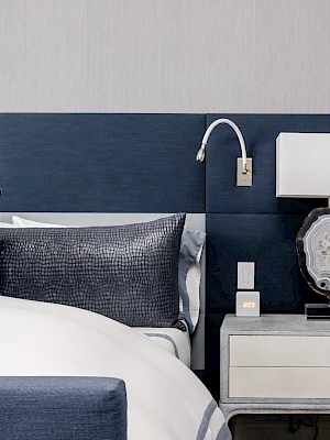 The image shows a modern bedroom with a blue and white color scheme, featuring a bed, headboard, pillows, a nightstand, lamp, and decor.