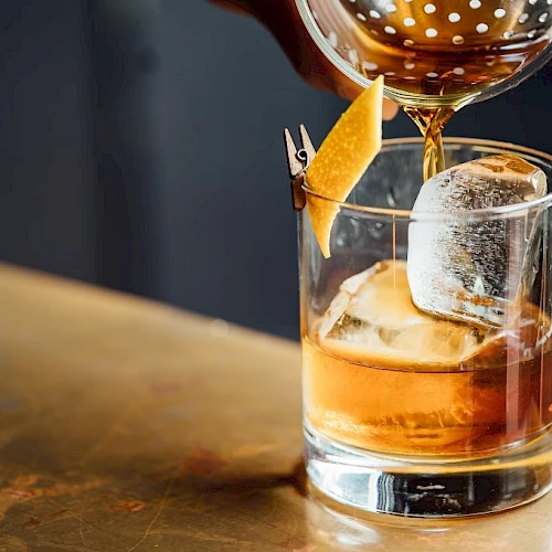 A drink is being poured into a glass with a large ice cube and an orange twist, on a bar counter.
