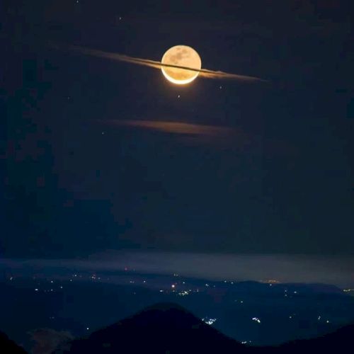 A full moon appears with a cloud forming a ring around it, resembling Saturn. The scene is over a nighttime landscape with scattered lights.