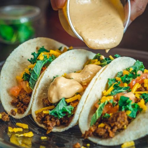 Three tacos filled with seasoned meat, cheese, and lettuce are on a plate. Sauce is being poured over them from a small container.