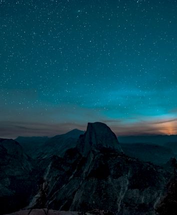 A starry night sky over a mountainous landscape, with a prominent peak silhouetted against the horizon and a glow from the setting sun.