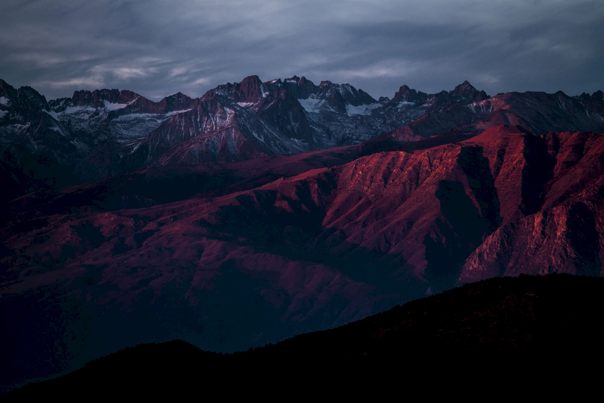 A mountain range with peaks partially covered in snow, bathed in a dramatic red and purple light, under a moody sky.