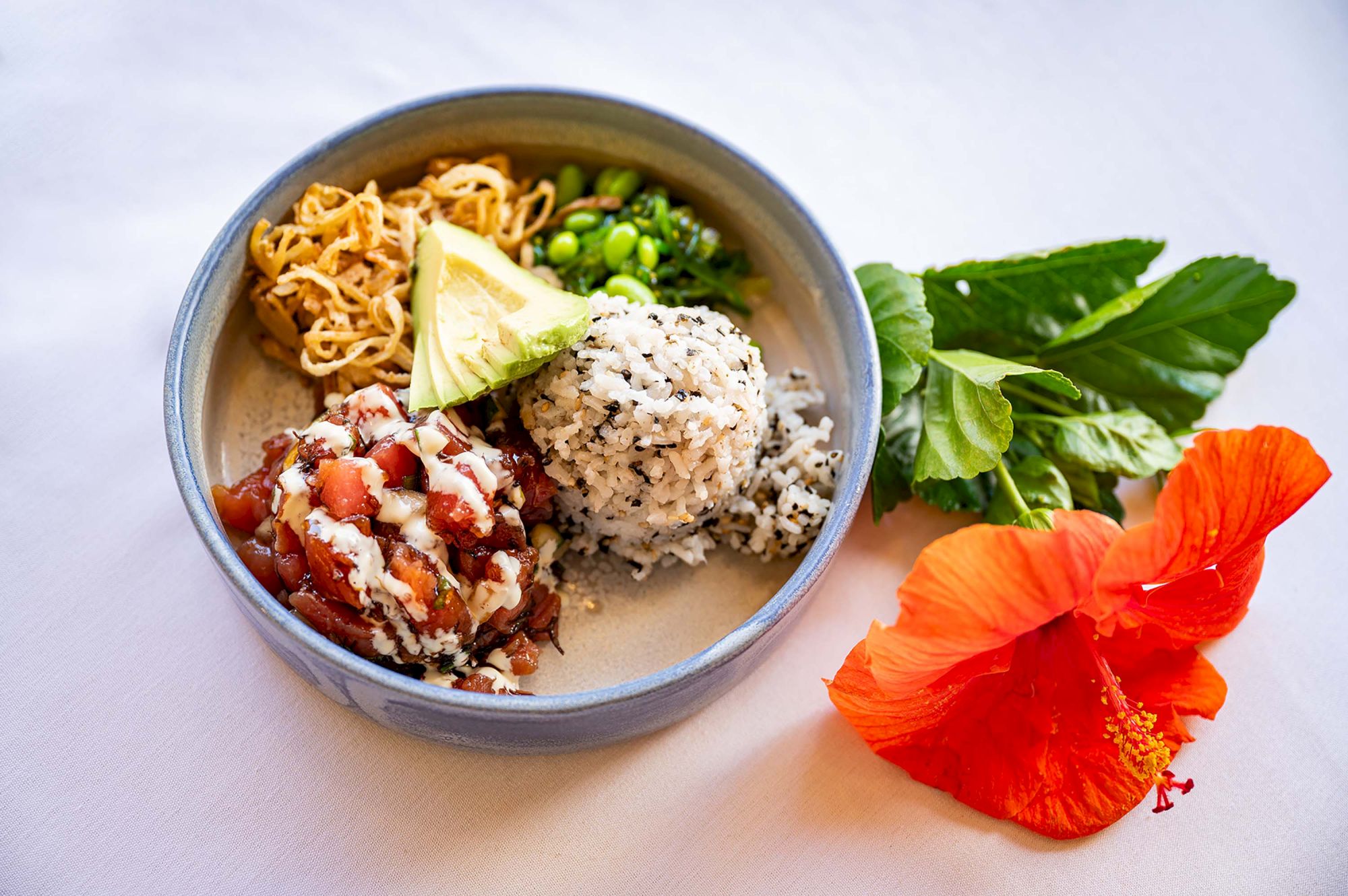 A bowl with noodles, avocado, mixed rice, edamame, and a tomato-based dish beside a hibiscus flower and greens on a white surface.
