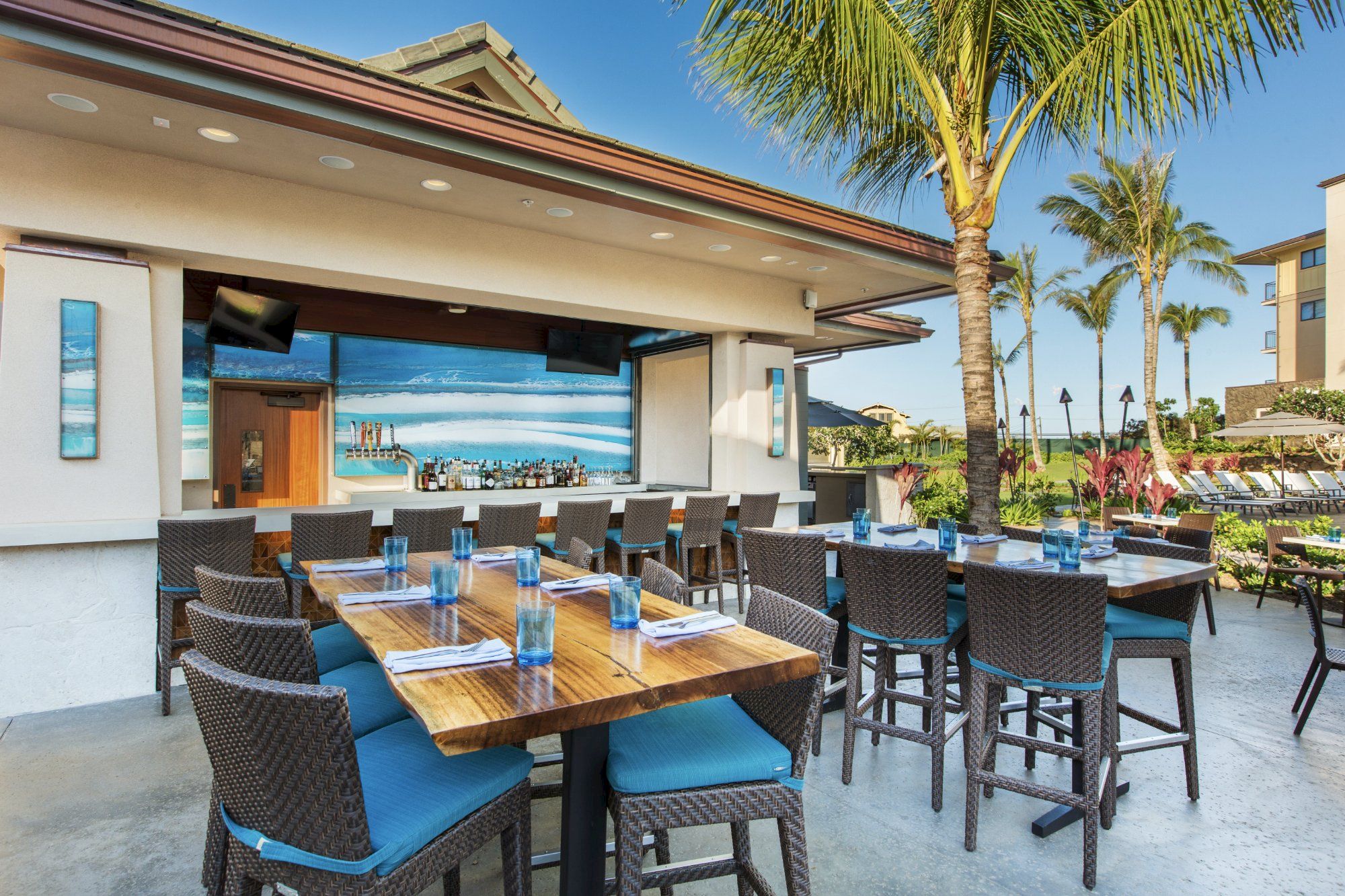 The image shows an outdoor dining area with wooden tables, chairs, and a bar, surrounded by palm trees under a clear blue sky.