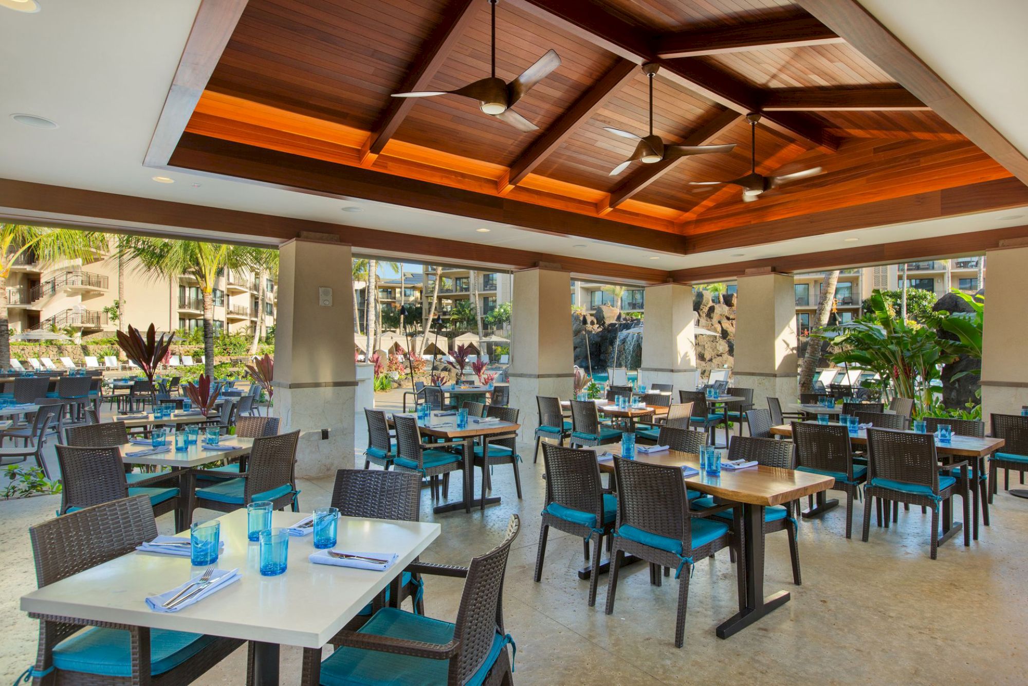 An open-air restaurant with wooden ceiling, modern tables and chairs, blue accents, overlooking a pool area with tropical plants.