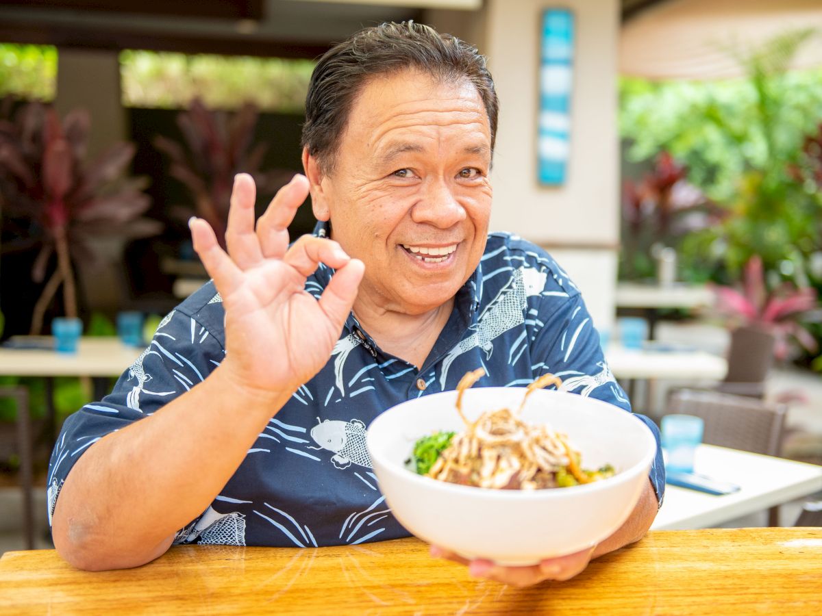 A person smiles, holding a bowl of noodles and making an "OK" gesture, sitting outdoors with greenery in the background.