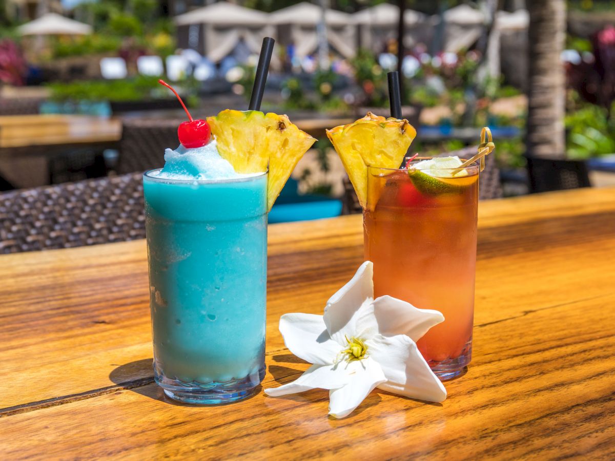 Two tropical cocktails garnished with fruit on a wooden table, with white flowers nearby, in an outdoor setting.