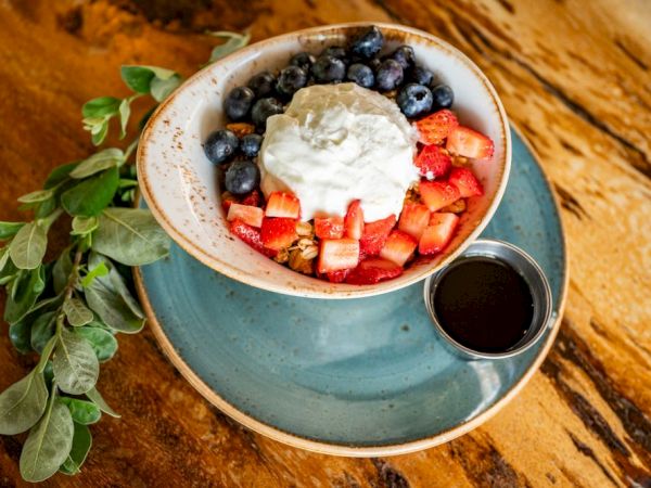 A bowl of yogurt topped with blueberries, strawberries, and granola, accompanied by a small cup of syrup on the side.