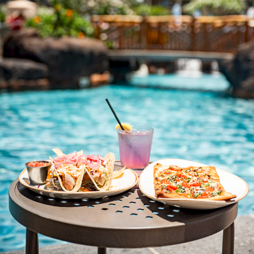 A table by a pool with tacos, pizza, and a purple drink. Palm trees and a bridge are visible in the background.