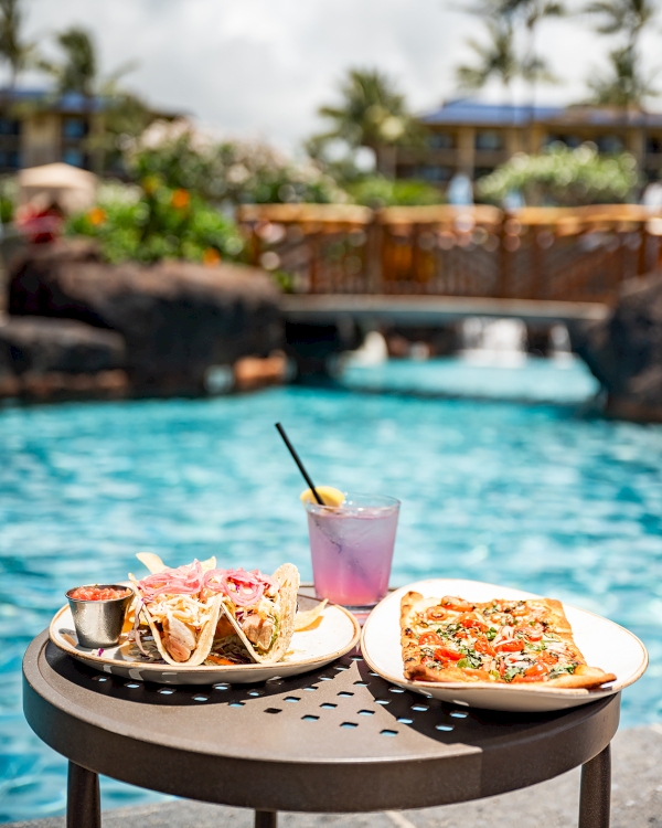 A table by a pool with tacos, pizza, and a purple drink. Palm trees and a bridge are visible in the background.