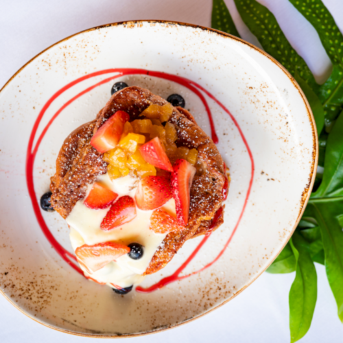 A plate of French toast topped with cream, sliced strawberries, diced fruit, and blueberries, with red sauce drizzle, next to green leaves.