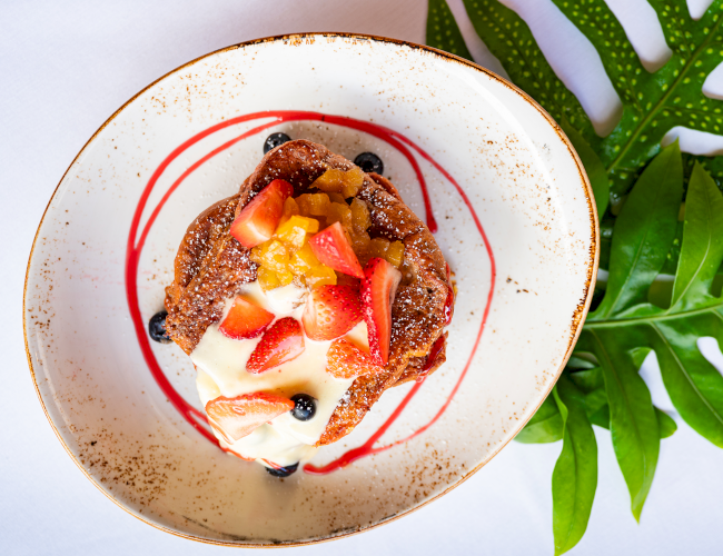 A plate of French toast topped with cream, sliced strawberries, diced fruit, and blueberries, with red sauce drizzle, next to green leaves.