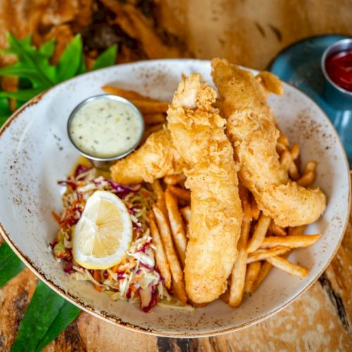 The image shows a plate of fish and chips with tartar sauce, coleslaw, and a lemon wedge, served with ketchup on the side.
