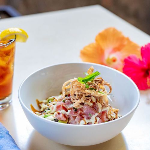 A bowl of salad with crispy toppings, a glass of iced tea with lemon, a napkin, and two hibiscus flowers on a table.