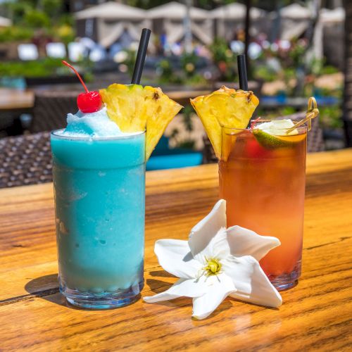 Two tropical cocktails with fruit garnishes and a white flower on a wooden table outdoors.