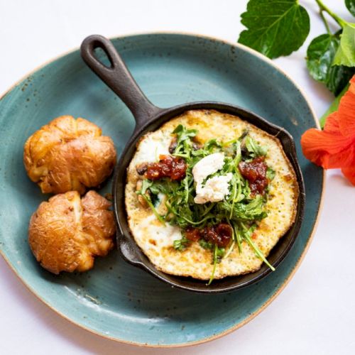 A skillet with a frittata topped with greens and cheese, served with two bread rolls on a plate, next to a hibiscus flower.