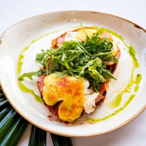 A plate with an open-faced sandwich, topped with arugula, an egg, and a drizzle of green sauce, on a white plate with a leaf garnish.