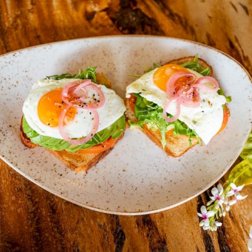 Two pieces of toast topped with fried eggs, sliced red onions, and greens on a white oval plate. Flowers are beside the plate.