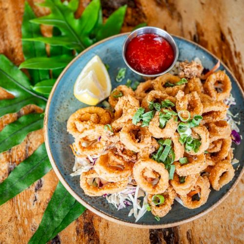 A plate of fried calamari with a lemon wedge, green garnish, and a side of red sauce on a wooden table.