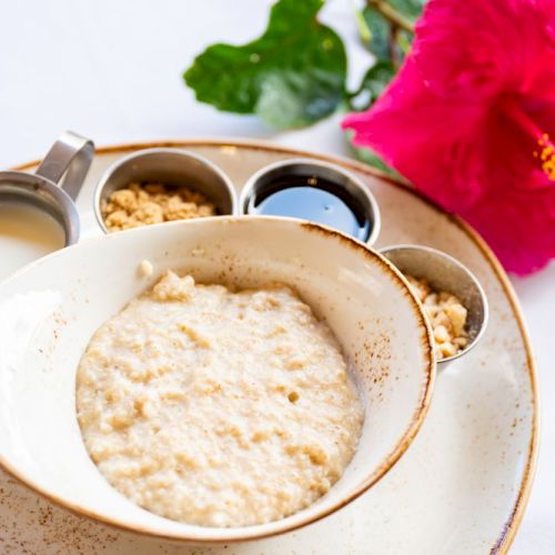 A bowl of porridge with cream, nuts, syrup, and a red flower beside it on a plate.
