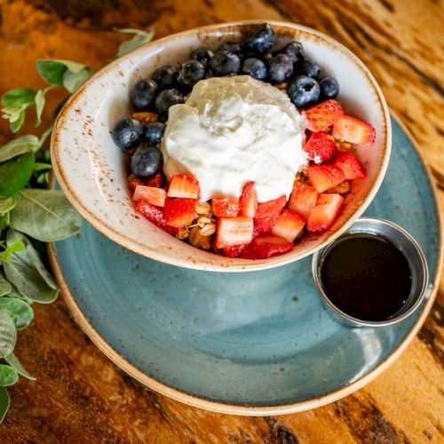 A bowl with yogurt, blueberries, strawberries, and granola, with a small cup of syrup on a blue plate, garnished with green leaves.