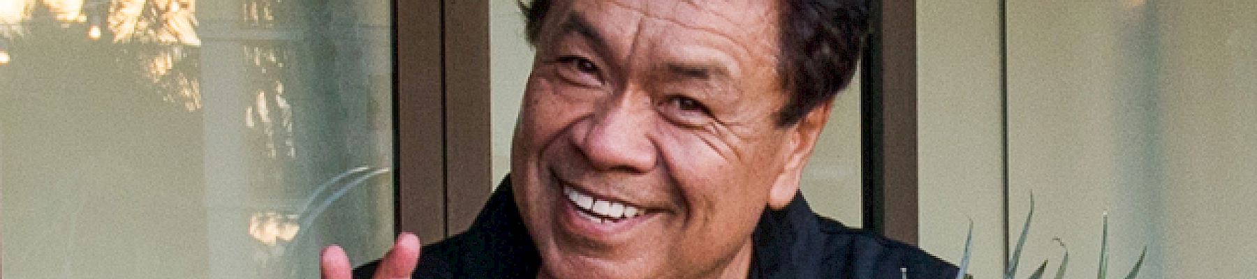 A person smiling and holding a pineapple while making an "OK" hand gesture, standing in front of a window with trees visible outside.