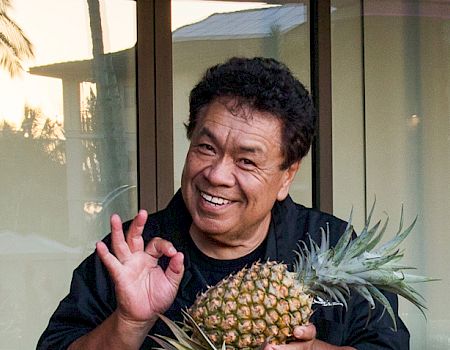 A person smiling and holding a pineapple while making an "OK" hand gesture, standing in front of a window with trees visible outside.