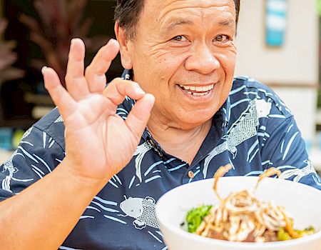 A man in a patterned shirt holds a bowl of food, smiling and making an "OK" gesture with his hand.