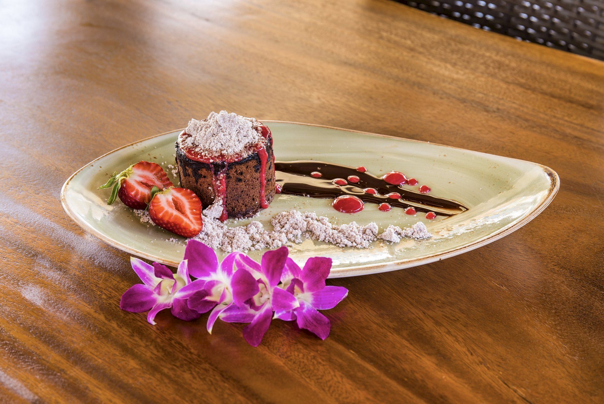 A dessert with strawberries, powdered topping, chocolate sauce, and purple flowers on a uniquely shaped plate.