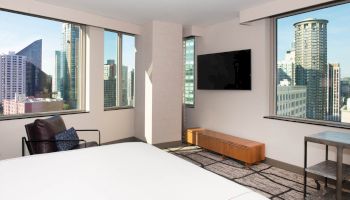 A modern hotel room with large windows showing city views, a bed, TV, chair, table, and bench against a neutral color scheme.