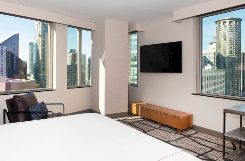 A modern hotel room with large windows showing city views, a bed, TV, chair, table, and bench against a neutral color scheme.