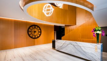 A modern lobby features a marble reception desk, wooden walls, a large clock, elegant lighting, and a vase of flowers for decoration.