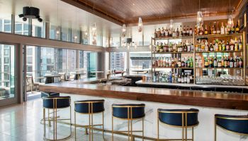 A modern bar with high stools, a variety of bottles on display, and large windows overlooking a city view in a sleek, stylish setting.