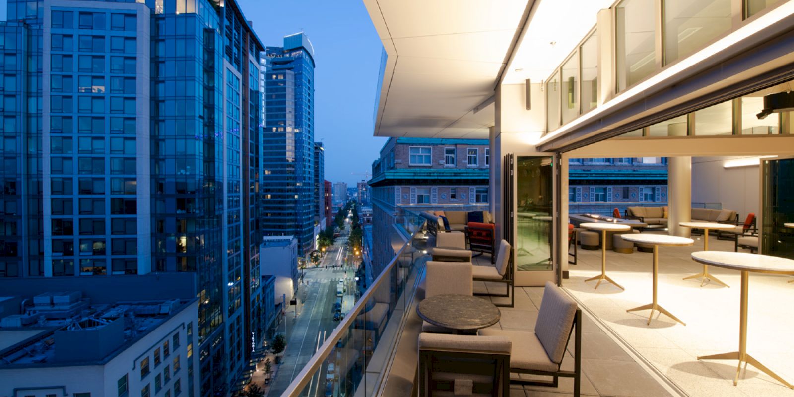 The image shows a modern cityscape with tall buildings and an elegant terrace with seating, illuminated in the evening.