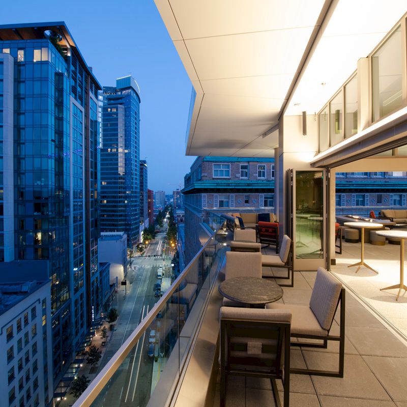 A modern rooftop terrace at dusk overlooks a city street lined with tall buildings, featuring outdoor seating and a sleek design.