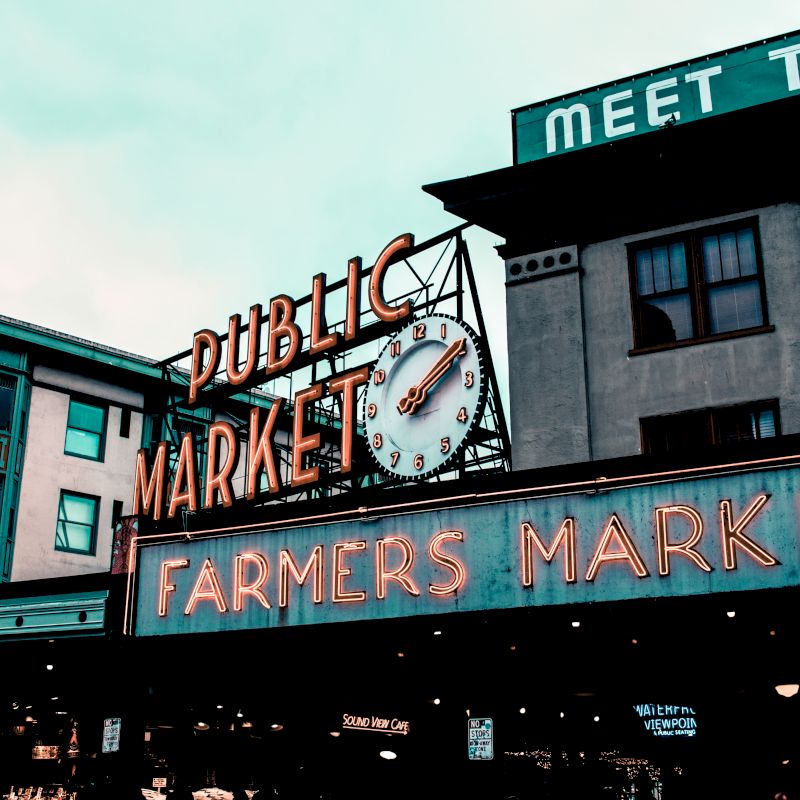 The image shows a famous public farmers market sign with neon lights and a clock, located in an urban area during the day.