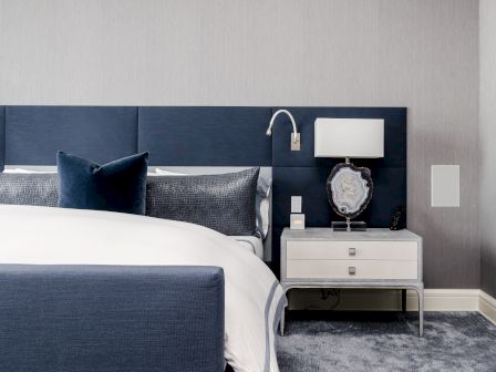 A modern bedroom with a white and navy color scheme, featuring a bed, blue headboard, nightstand, lamp, and decorative pillows.