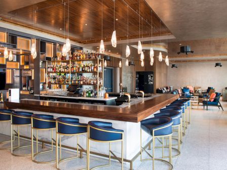 This image shows a modern bar with a central counter, blue stools, pendant lights, and shelves stocked with bottles in a trendy setting.