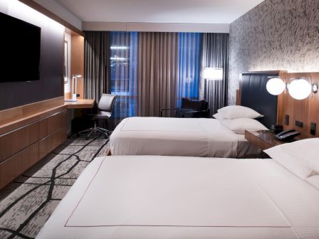 The image shows a modern hotel room with two beds, a wall-mounted TV, a work desk, and soft lighting.