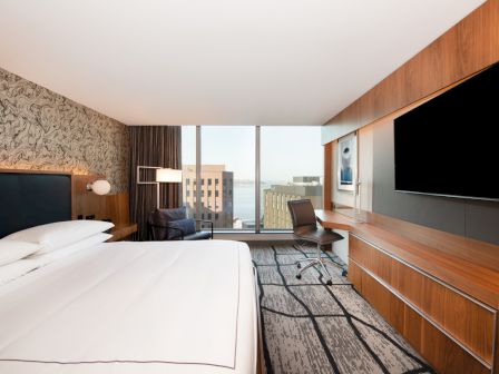 A modern hotel room with a large bed, flat-screen TV, desk, and city view through a window, featuring contemporary decor and furnishings.